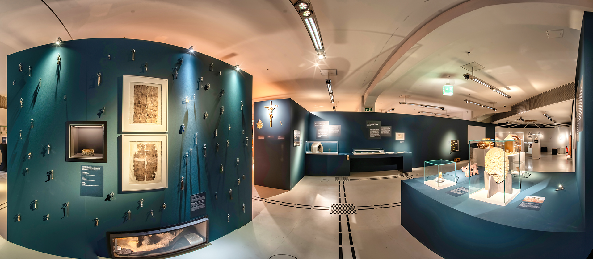 Das Bild zeigt einen blau gestrichenen Raum mit Vitrinen und Objekten an der Wand. Durch den Raum verläuft ein Bodenleitsystem. Ganz links ist eine Wand mit ganz vielen Schlüsseln. 