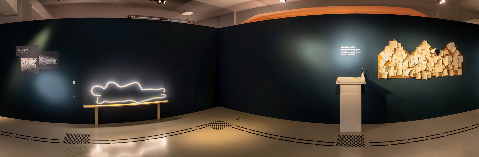 Das Bild zeigt eine dunkle Wand mit einem Bodenleitsystem davor. Auf der linken Seite ist eine Leuchtskultpur an der Wand befestigt. Sie zeigt die Umrisse einer schlafenden Person auf einer Holzbank. Rechts sind drei Pinnwände in Haus-Form mit vielen Notizzetteln daran.